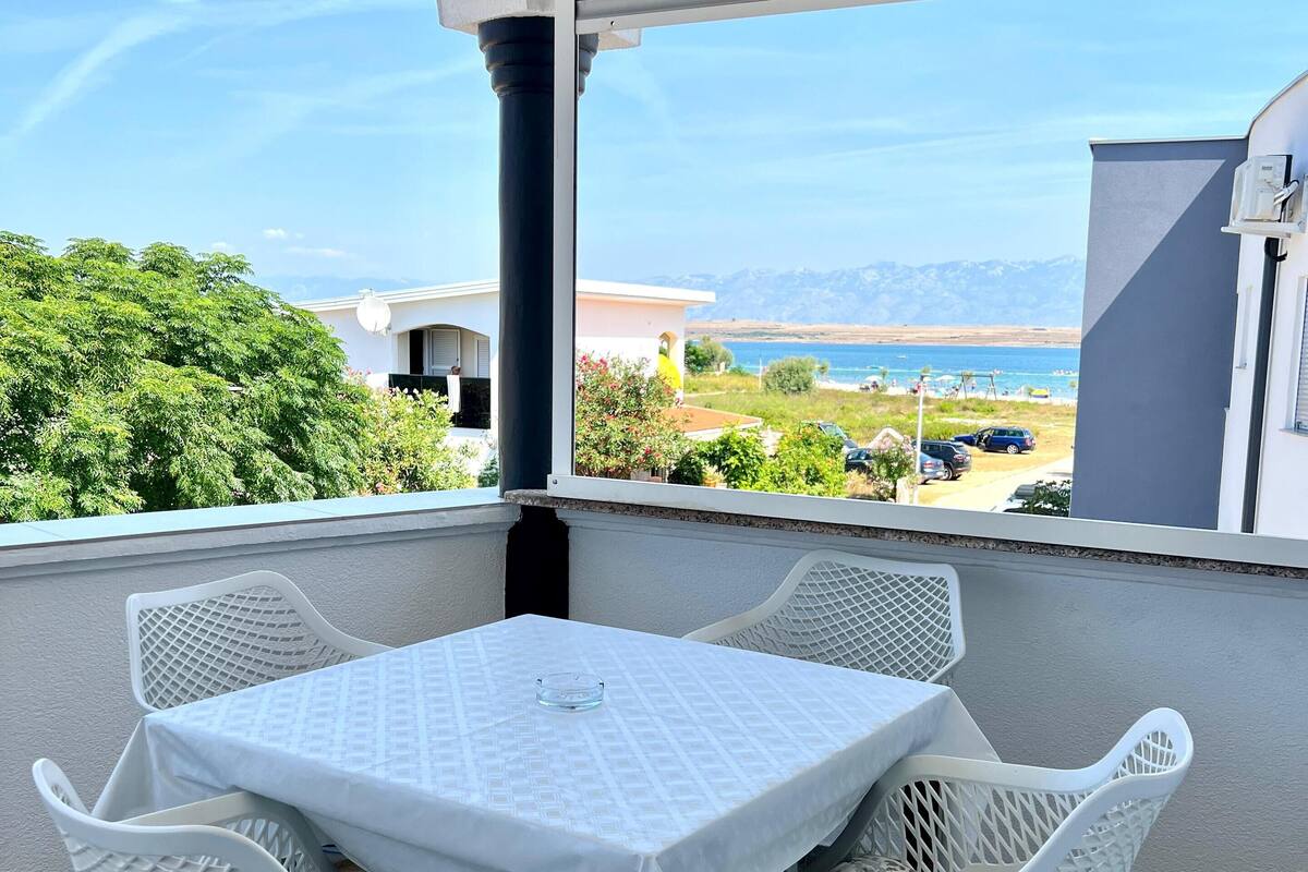 A balcony with a table and chairs, overlooking a beach and mountains.