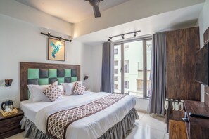 A modern hotel room with a large bed, a wooden headboard, a flat-screen TV, a desk with a remote control, and a view of the outside through a sliding glass door. - EL CLASSICO (Rajkot)