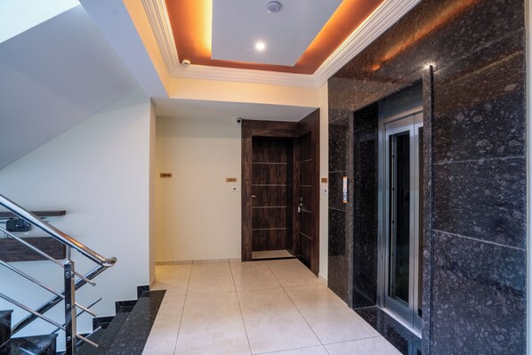 A modern hallway with a marble floor, a wooden door, and a glass elevator. - EL CLASSICO (Rajkot)