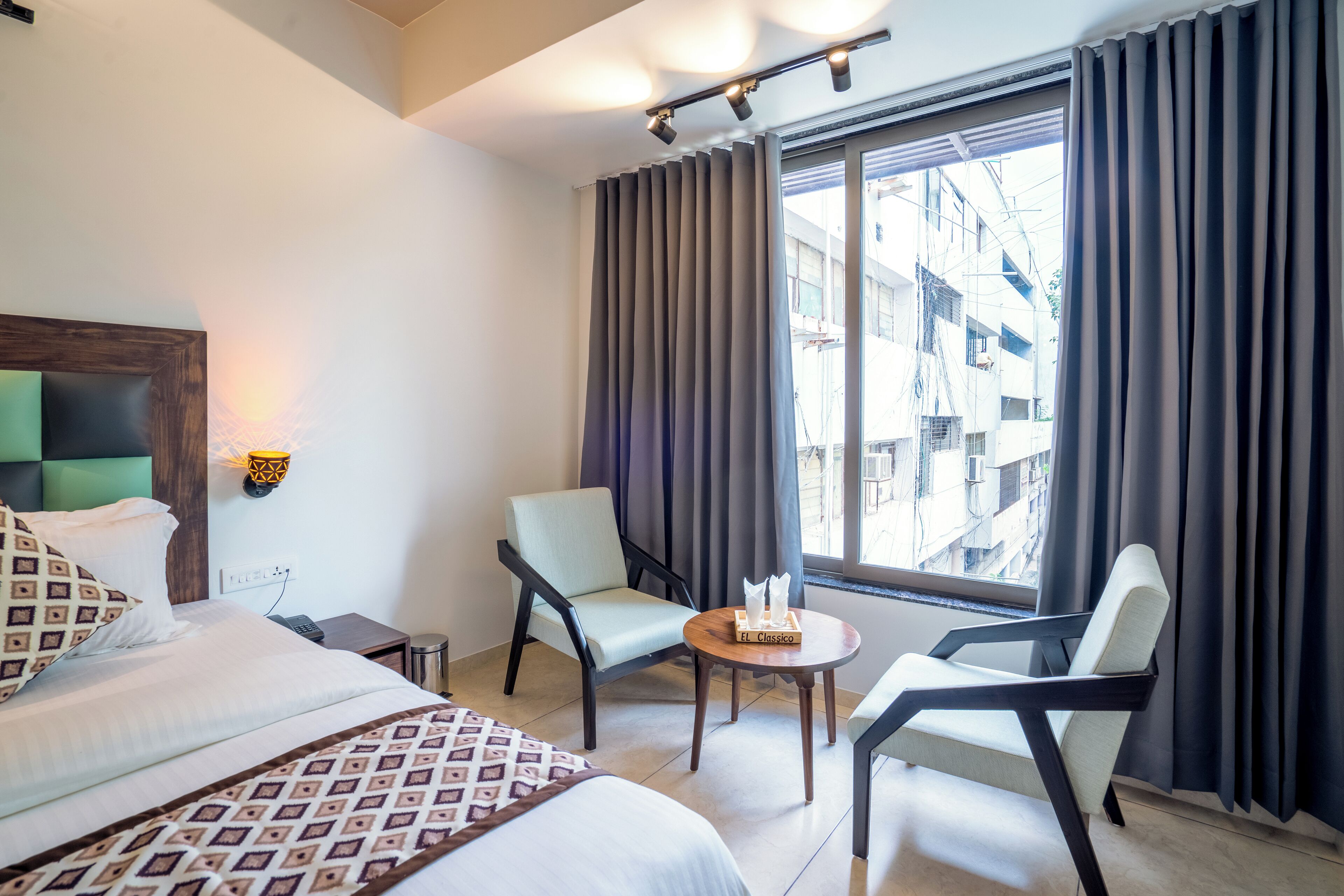 Quarto de hotel com cama, duas cadeiras, uma mesinha, abajur e janela com cortinas.