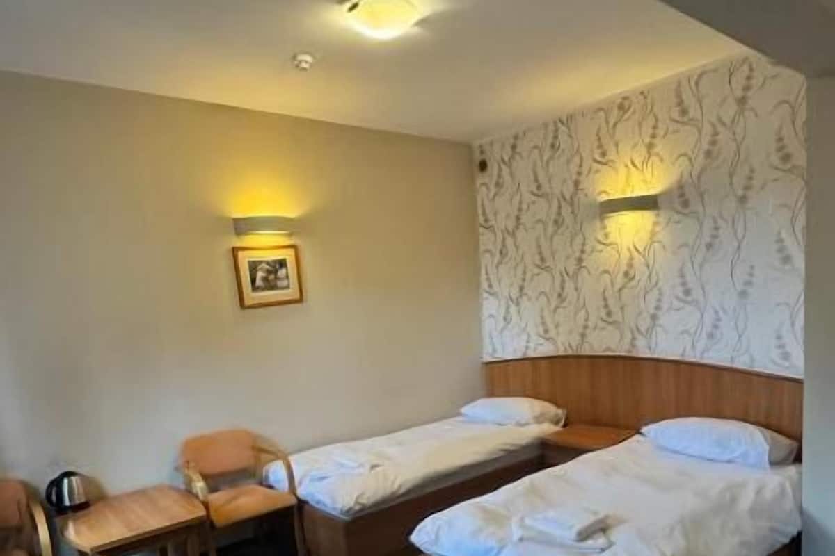 Ein Hotelzimmer mit zwei Einzelbetten, einem Holzkopfteil, einem kleinen Tisch mit einem Stuhl, einer Wandlampe und einem gerahmten Bild an der Wand.