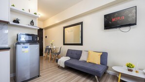 A modern living room with a grey sofa, a small table with a vase, a microwave, and a refrigerator. A TV mounted on the wall displays the Netflix logo. - Queen Studio, City View - Virgo Nest Rentals (Cebu City)
