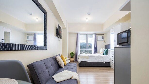 A modern hotel room with a sofa, a bed, a TV, and a large window with curtains. - Queen Studio, City View - Virgo Nest Rentals (Cebu City)