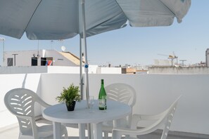 Une table ronde blanche, deux chaises, une plante en pot, une bouteille et un verre, le tout sur une terrasse sur le toit.
