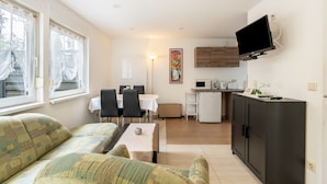 A compact living space with a sofa, dining table, chairs, a kitchen area with a microwave, and a TV mounted on the wall.