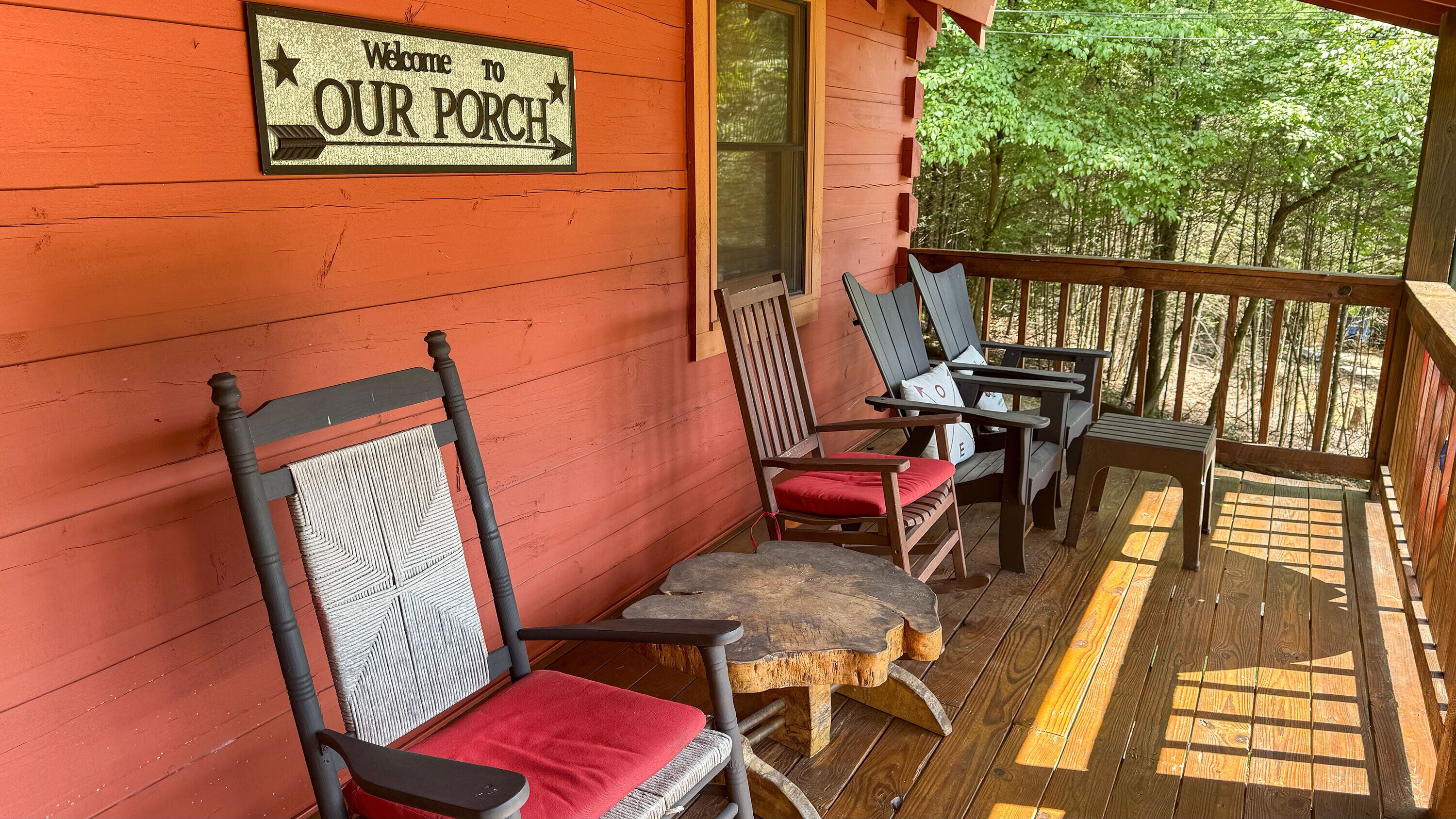A porch with a 'Welcome to OUR PORCH' sign, wooden chairs, a small table, and a view of trees through the window.