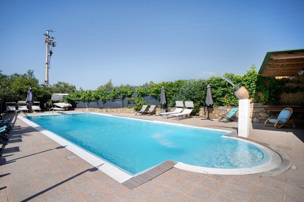Una piscina rectangular con borde de baldosas, rodeada de sillas de descanso y sombrillas.