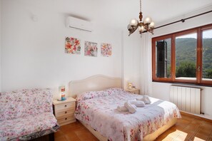 Un dormitorio con dos camas, una ventana con vistas a las colinas, una lámpara en el techo y aire acondicionado instalado en la pared.