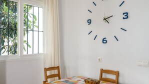 A room with a wall clock, a table with a patterned tablecloth, two wooden chairs, and a window with a view of greenery. - Apartment 'Atardecer En Conil 2' with Air Conditioning (Conil de la Frontera)