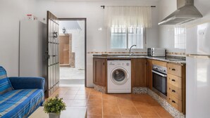A compact kitchen with a washing machine, oven, and a dining area with a blue sofa. - Apartment 'Atardecer En Conil 2' with Air Conditioning (Conil de la Frontera)