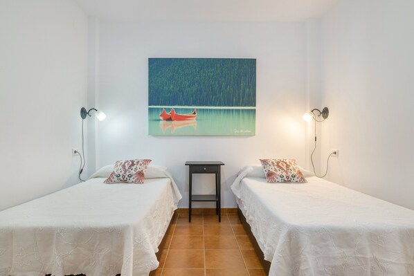 Une chambre avec deux lits simples, une petite table, un tableau représentant un lac avec un bateau, et deux lampes fixées au mur.