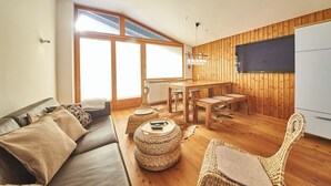 A cozy living room with a sofa, coffee table, and wicker chairs. A dining area with a table and chairs is adjacent to the living room. The room has wooden walls and a large window providing natural light. - SunnySide Appartements (Hinterglemm)