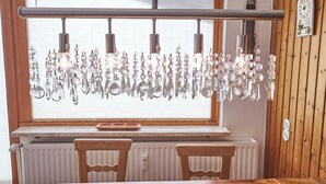 A dining area with a wooden table and chairs, a window with blinds, and a hanging crystal chandelier. - SunnySide Appartements (Hinterglemm)
