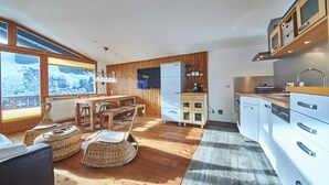 A modern kitchen with wooden cabinetry, a dining area with a wooden table and chairs, and large windows with a snowy view. - SunnySide Appartements (Hinterglemm)
