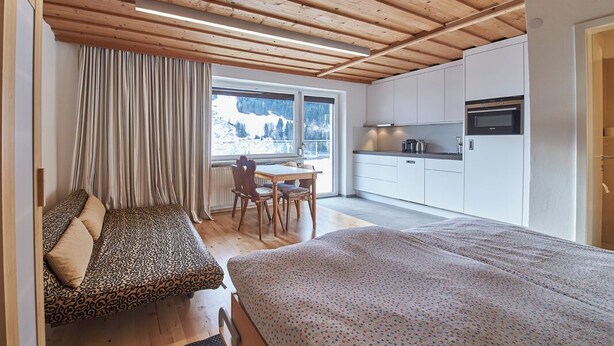 A cozy room with a wooden ceiling, a bed, a sofa, a dining table with chairs, and a kitchen area with cabinets and appliances. - SunnySide Appartements (Hinterglemm)