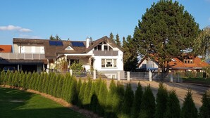 Una casa de dos pisos con techo inclinado, paneles solares y un gran árbol en el jardín.