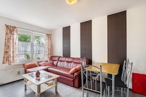 Una sala de estar con un sofá de cuero, una mesa de centro de vidrio, una mesa de comedor de metal con sillas y una ventana con cortinas estampadas.