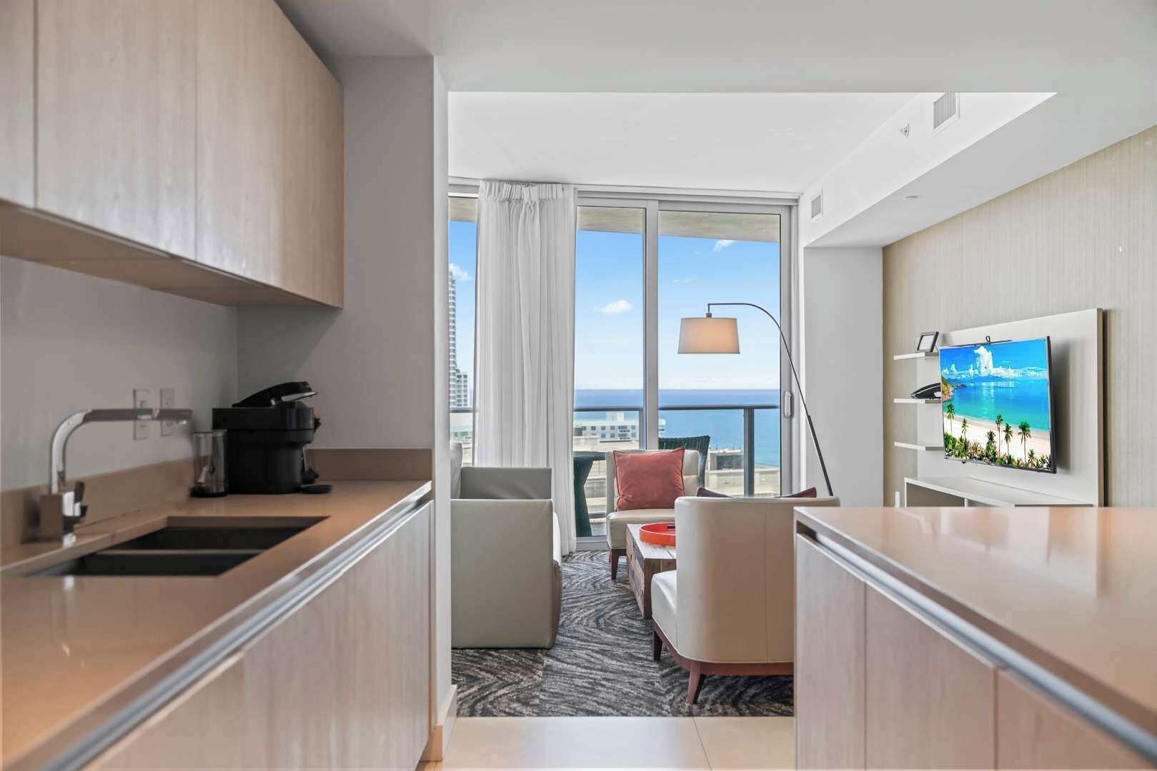 Una habitación de hotel moderna con kitchenette, televisor de pantalla plana, balcón con vista al océano y un amplio ventanal con cortinas.