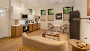 A modern kitchen with wooden cabinets, a built-in microwave, and a coffee maker. There is a dining area with a round table and two chairs. A sofa and a coffee table are in the living room.