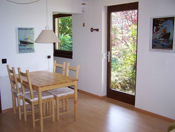 A dining area with a wooden table and chairs, a window with a view of greenery, a door with a key, and a wall-mounted lamp.