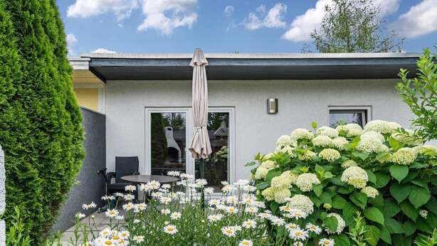 A modern building with a patio area featuring a table and chairs, surrounded by white flowers and greenery. - Holiday Home 'Bungalow 73' with Private Terrace and Private Garden (Zadelsdorf)