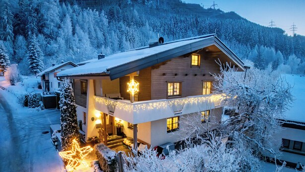 Un chalet à deux étages, doté d’un toit en pente, entouré d’arbres couverts de neige et avec une chaîne de montagnes en toile de fond.