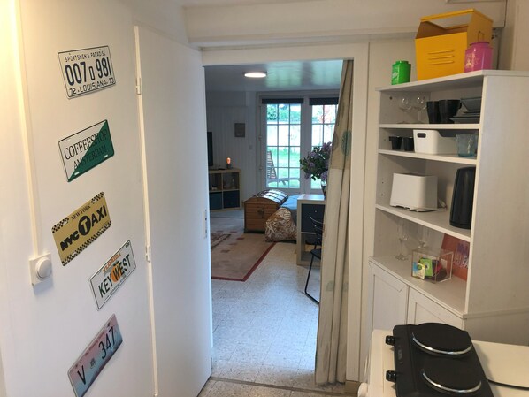 A hallway with a door leading to a living room. The hallway has a white door with several stickers, including a sportsmen's paradise license plate and a NYC taxi sticker. The living room has a sofa, a coffee table, and a window with curtains. There is a white cabinet with various items on top, including a green container and a pink container.