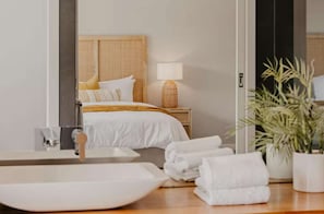Una habitación de hotel moderna con una cama grande, una mesita de noche con lámpara, un baño con lavamanos amplio y una planta en maceta.