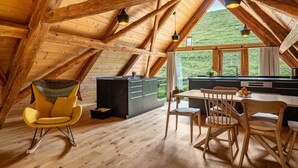 Une pièce aux hauts plafonds et aux poutres apparentes, avec une table à manger et des chaises, un coin cuisine et une vue sur une colline verdoyante.