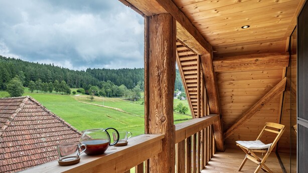 Un balcon en bois offrant une vue sur un paysage verdoyant, avec une chaise et une table sur laquelle se trouvent une théière et une tasse.