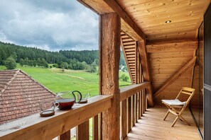 A wooden balcony with a view of a green landscape, a chair, and a table with a teapot and cup.