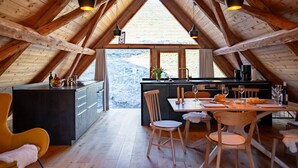 Une pièce aménagée en style mansarde, comprenant une cuisine et un coin repas, avec des poutres apparentes, une grande fenêtre et une vue sur l’extérieur.