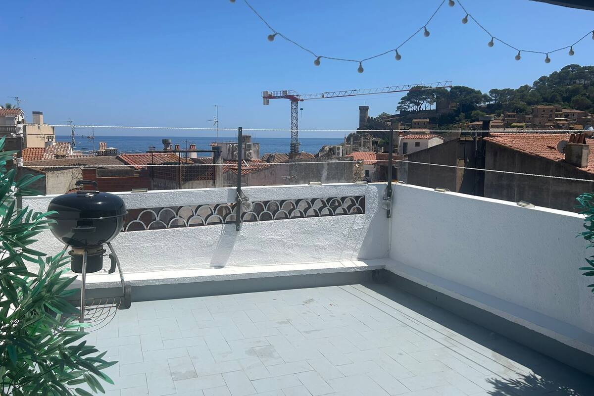A rooftop terrace with a barbecue grill, string lights, and a view of the sea and buildings.