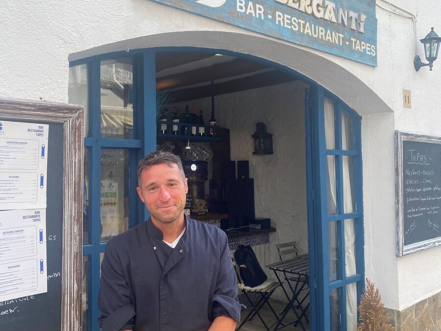 A man standing in front of a building with a sign that reads "EL BERGANT BAR RESTAURANT TAPES".
