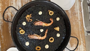 A top-down view of a dining table with a black pan containing seafood, white plates, and blue glasses. - Hotel & Restaurante Bergantí (Tossa de Mar)