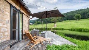Una terraza de madera con mobiliario exterior, un paraguas grande y vistas a un lago y un bosque.