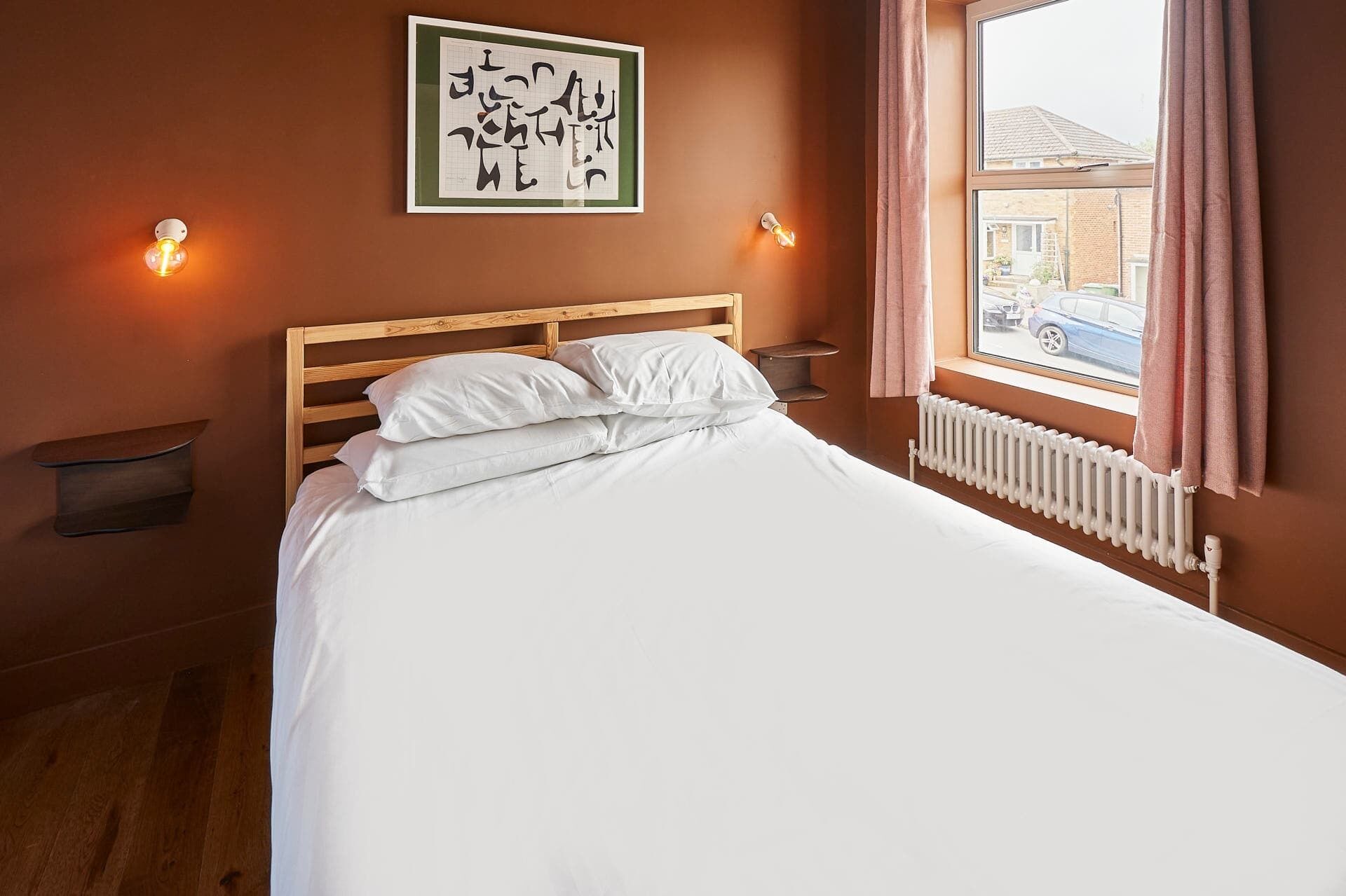 Un dormitorio con cabecera de madera, ropa de cama blanca, un radiador y vistas a la calle a través de una ventana con cortinas.