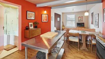 A modern kitchen with a central island, wooden dining table, and chairs. There is a built-in oven, a sink, and a window with blinds.