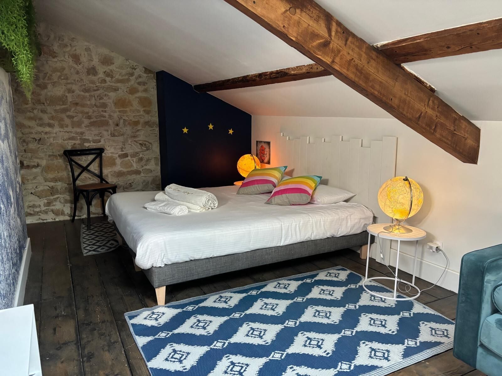 Un dormitorio con pared de piedra, cama con ropa de cama blanca, almohadas coloridas, una lámpara de globo y una alfombra estampada.