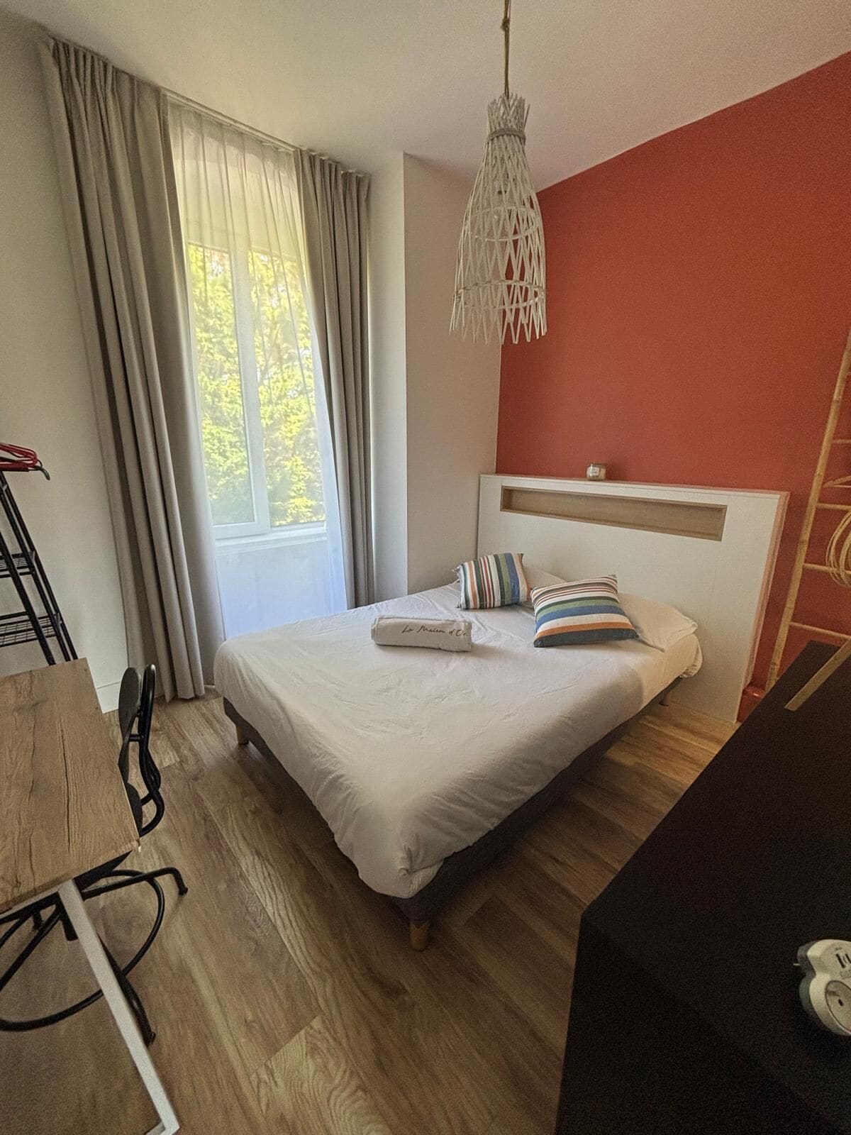 Une chambre à coucher avec un lit, un luminaire suspendu, une fenêtre avec des rideaux et un plancher en bois.
