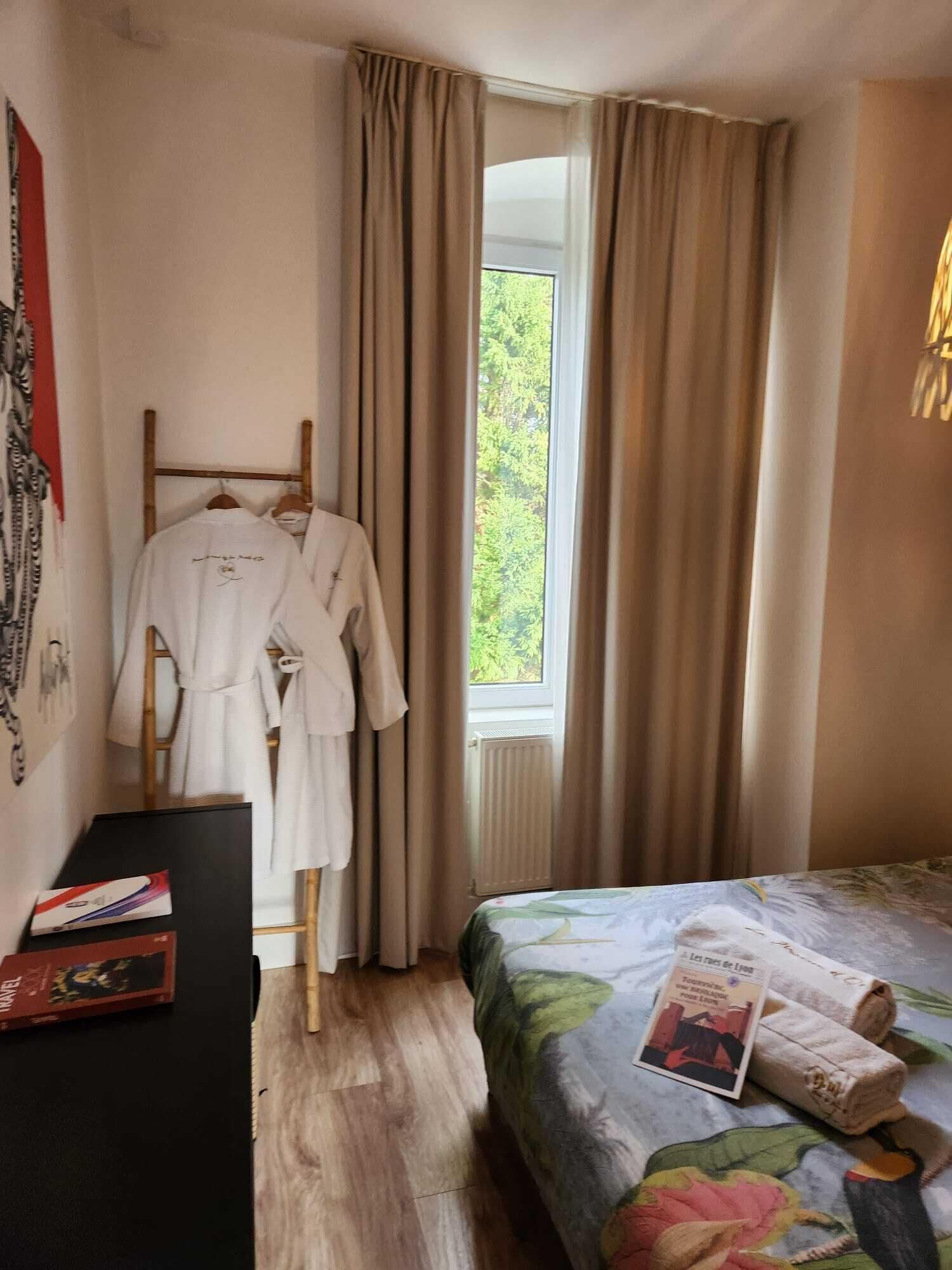 A bedroom with a bed, a window with curtains, a wooden rack with robes, and a small table with books.