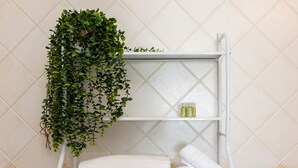 Une salle de bain avec un mur recouvert de carreaux blancs, une étagère blanche sur laquelle est posée une serviette blanche roulée, un petit flacon et une plante verte suspendue.