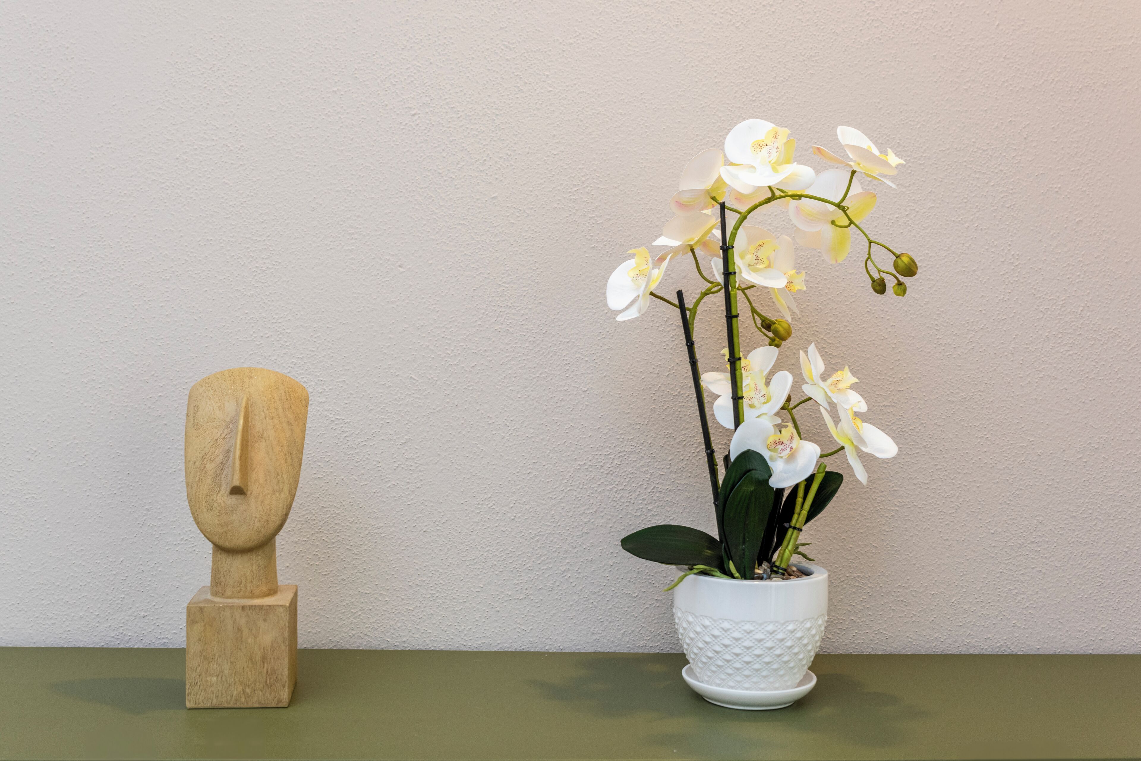 Un'orchidea in vaso accanto a una scultura in legno su un tavolo, con uno sfondo neutro.