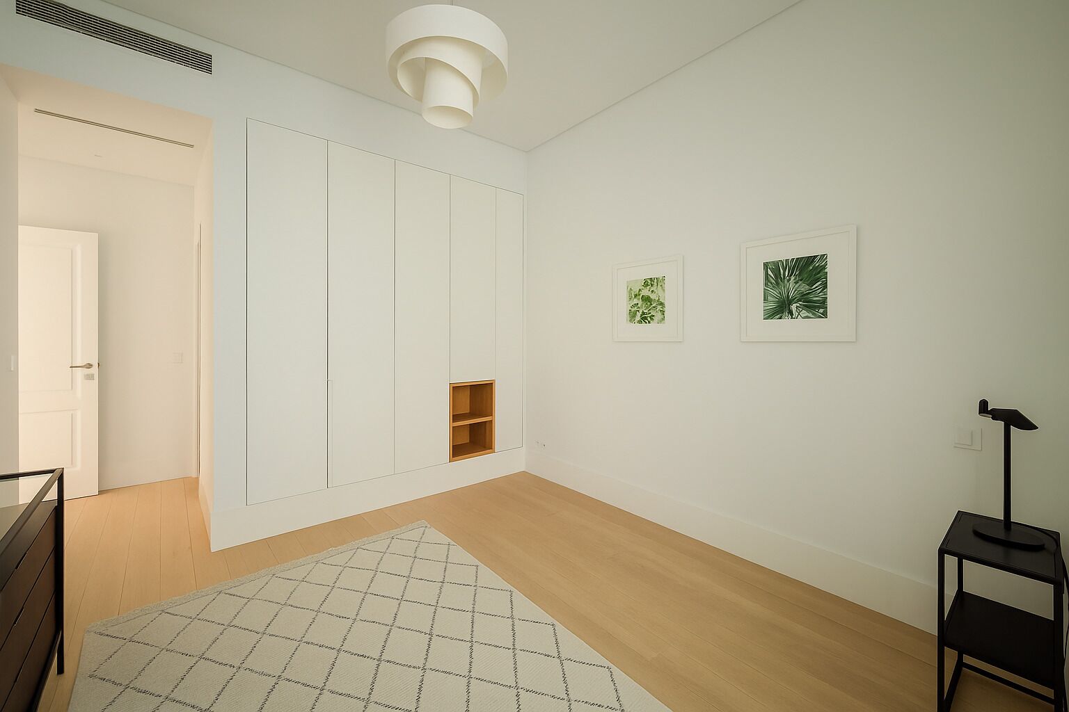 A modern, minimalist room with a wooden floor, a white door, a built-in wardrobe, a small shelf, and two framed botanical prints on the wall.