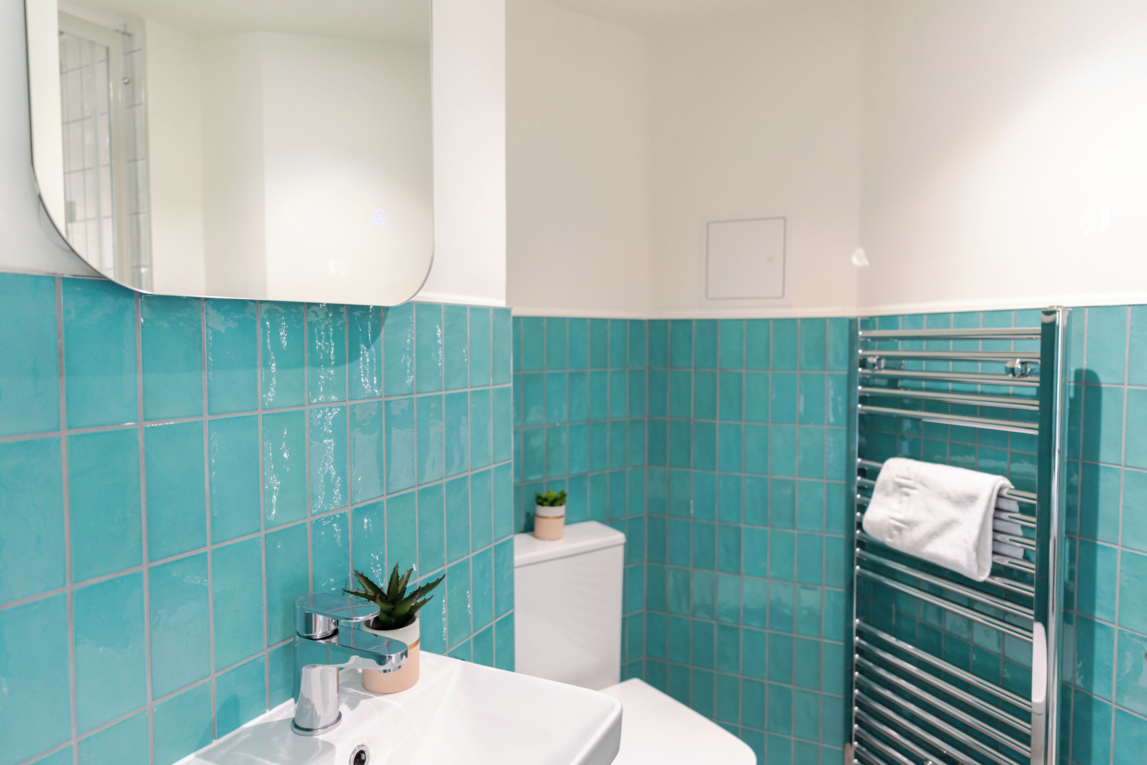 Un baño con azulejos turquesa, un lavabo blanco, un espejo, un perchero para toallas y una planta pequeña.