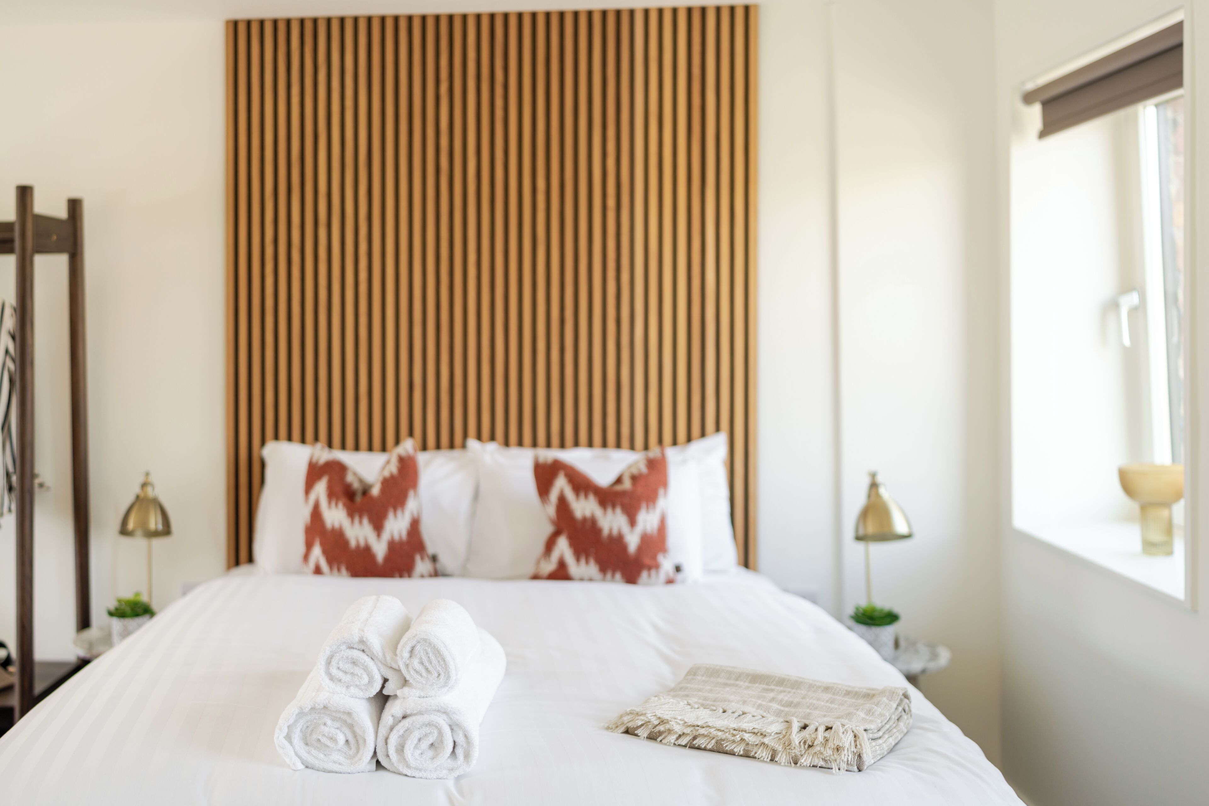 Un lit bien fait, avec des serviettes blanches pliées en rouleau et un plaid beige, une tête de lit en lattes de bois et deux lampes de chevet.