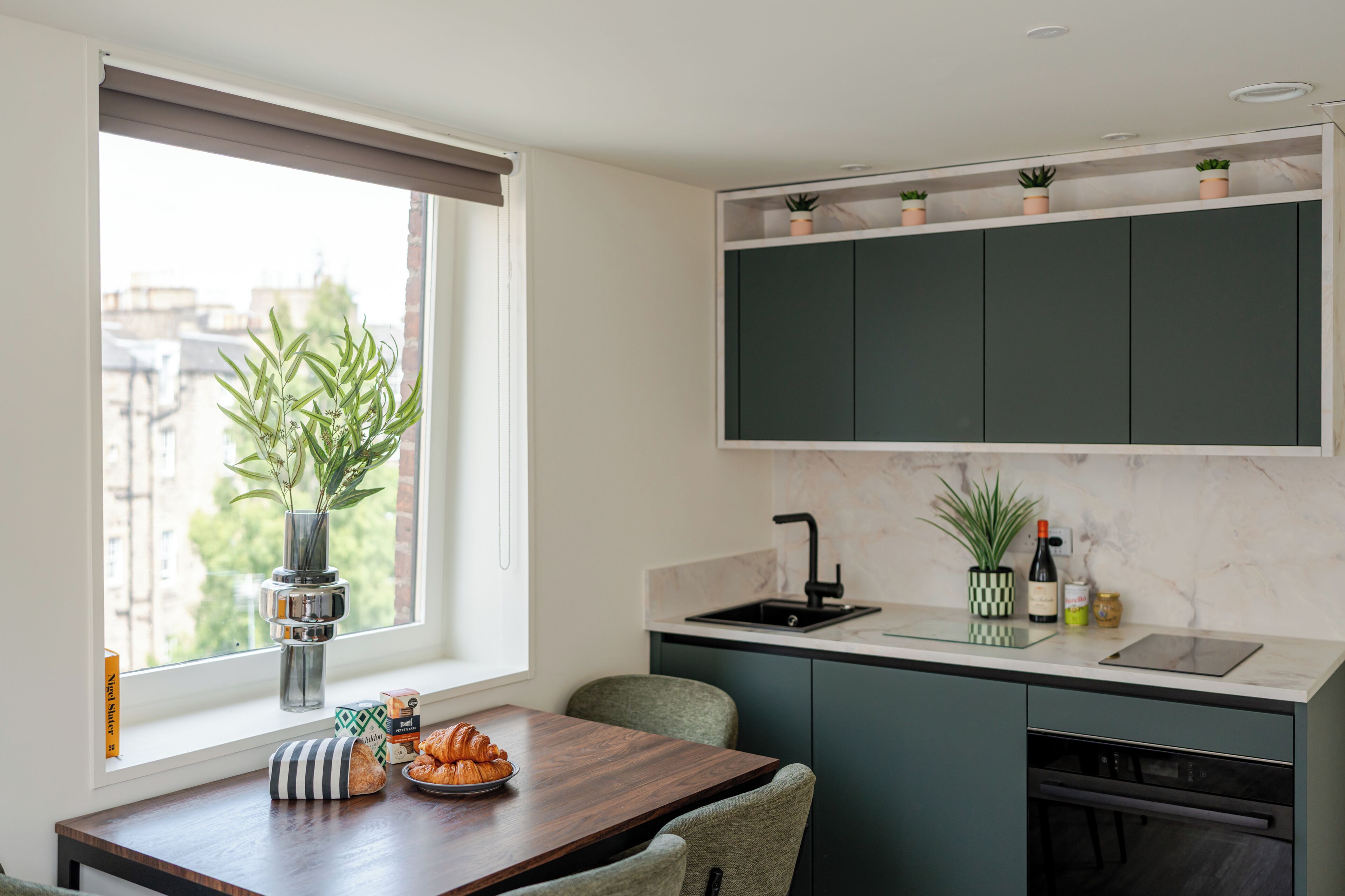 Et moderne køkken med mørkegrønne skabe, en marmor bordplade, et vindue med udsigt til bygninger, et spiseområde med bord og stole, og en potteplante på vindueskarmen.