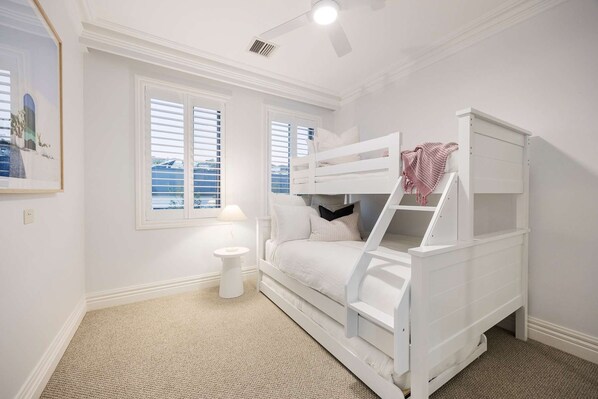 Un dormitorio con una cama nido, una mesita de noche pequeña, una lámpara y una ventana con persianas.