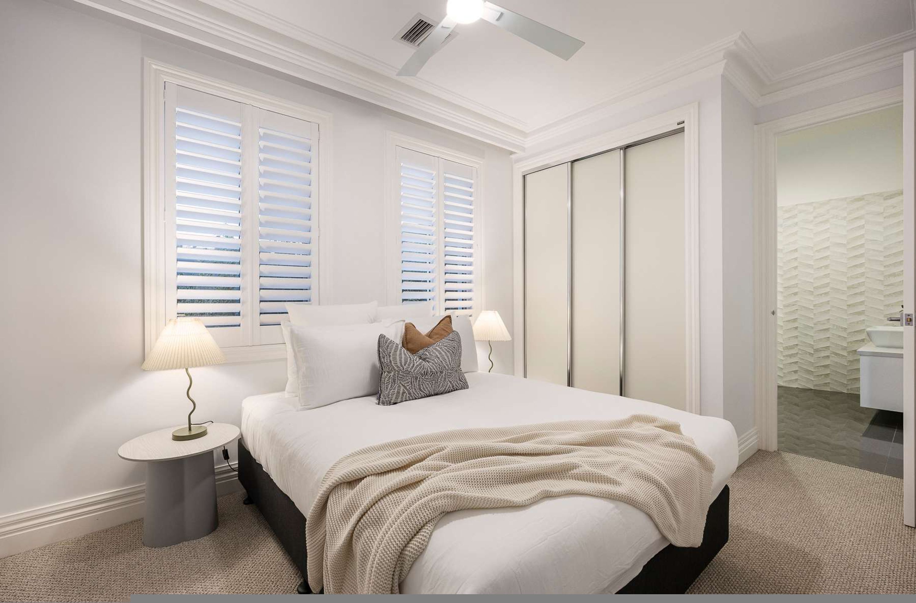 A bedroom with a bed, two bedside lamps, a nightstand, a closet, and a bathroom visible through an open door.