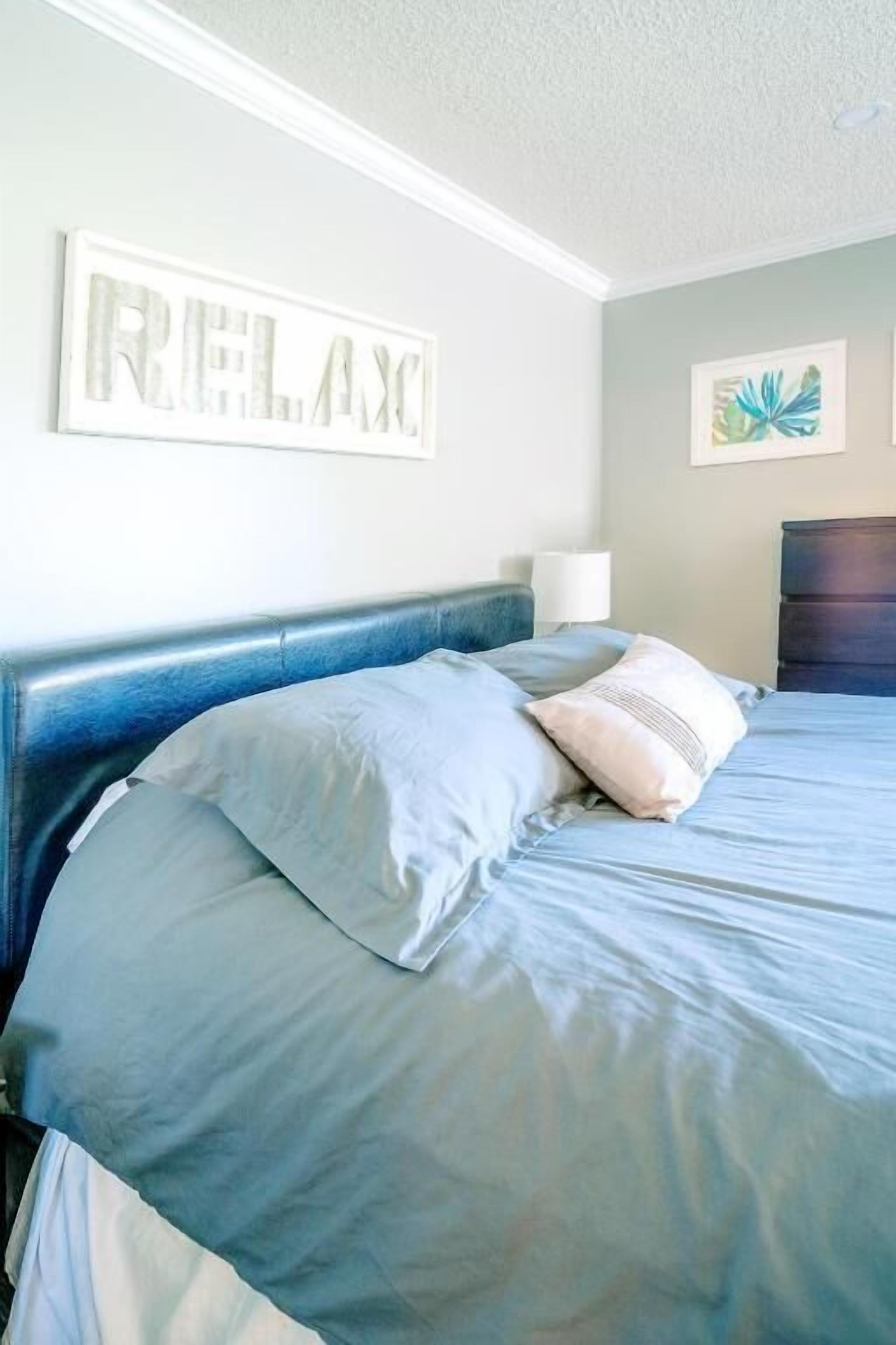 Ein Schlafzimmer mit Bett, Kissen, Kopfteil, einer Wandleuchte und einem gerahmten Kunstwerk.